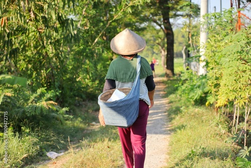 A woman wearing a caping or cone-shaped hat made of woven bamboo while carrying a basket made of cloth on her back, heading to the fields. photo