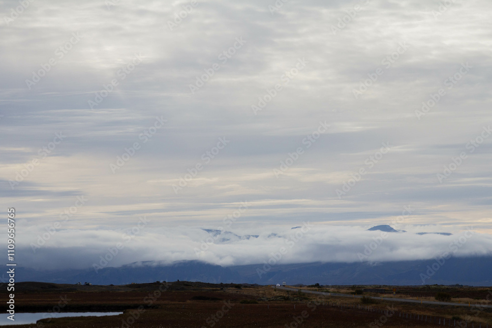 Fototapeta premium A winding road through a field, with towering, cloudy mountains looming in the distance, showcasing Iceland's raw beauty