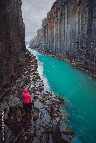 A female hiker in vibrant red outdoor gear exploring the basalt column formations and turquoise waters of Stuðlagil Canyon, Iceland.