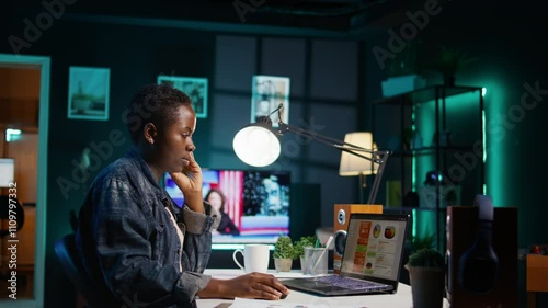 African american woman working from home, using laptop, looking at statistic charts. Remote worker in personal apartment office crosschecking important info between clipboard and notebook, camera B photo