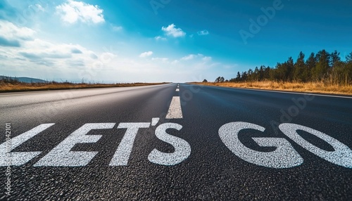 The Phrase Let'S Go Displayed On Highway In Middle Of Desolate Asphalt Road Beneath Stunning Blue Sky.
