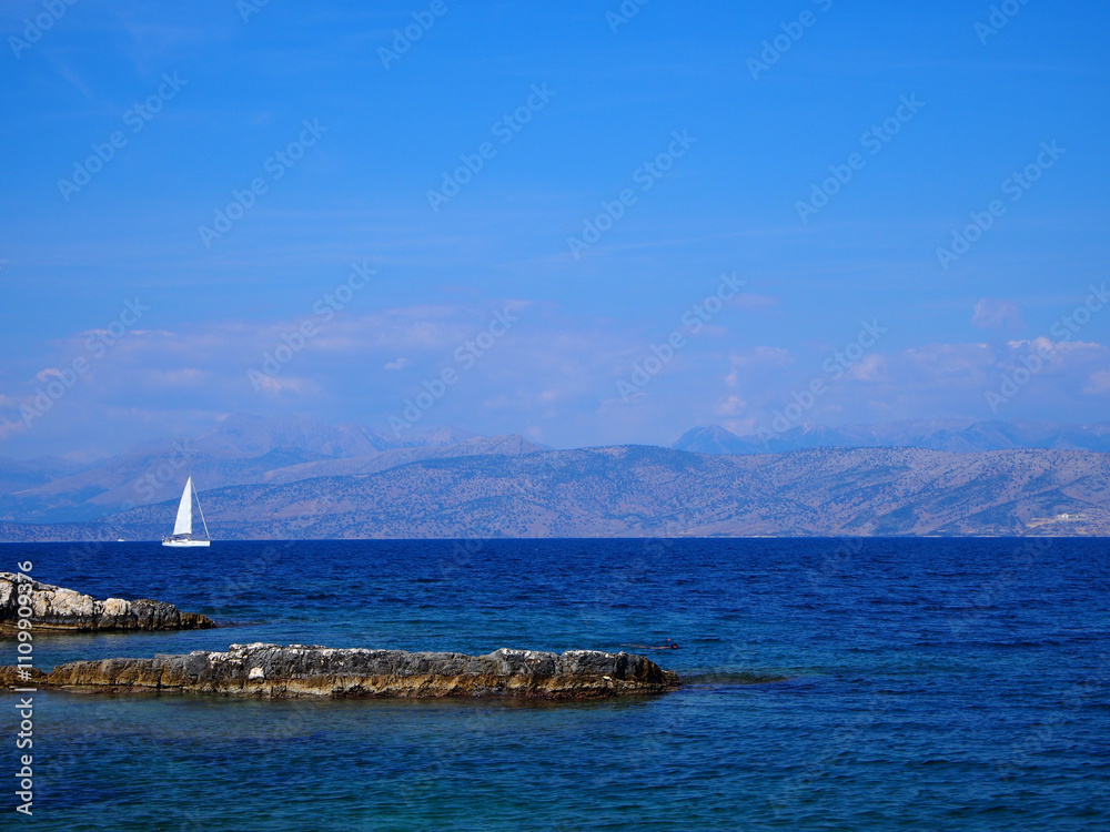 A single white sailboat in front of hills in the ionian sea in Corfu, Greece A single white sailboat in front of hills in the ionian sea in Corfu, Greece