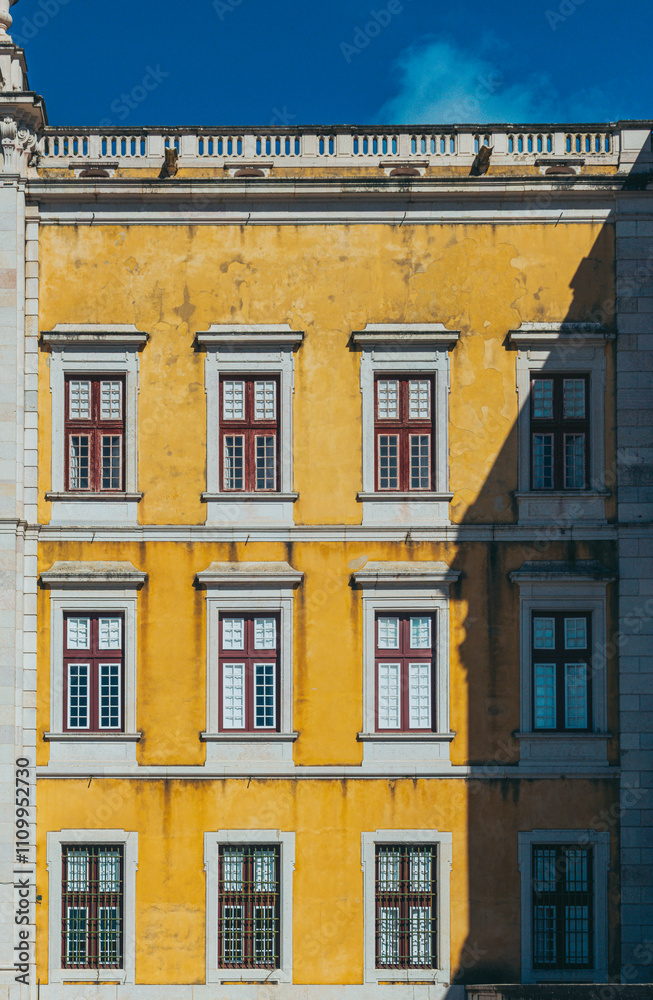 Fototapeta premium Sunlight is illuminating the yellow facade of mafra national palace in portugal, creating a captivating play of light and shadow on the windows and architectural details
