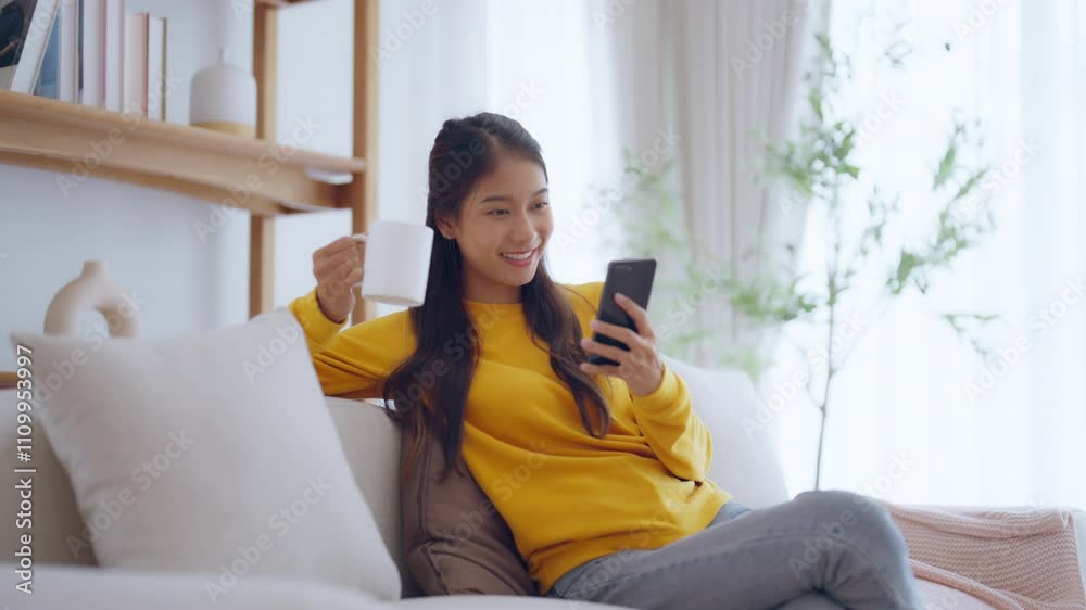 Happy young asian woman relax on comfortable couch at home texting messaging on smartphone, smiling girl use cellphone chatting, browse wireless internet on gadget, shopping online from home
