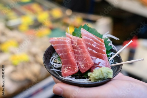 Fatty tuna sashimi slices glazed with soy sauce, and garnished with a perilla leaf and fresh wasabi, held in a woman’s hand at Tsukiji Outer Fish Market in Chuo, Tokyo, Japan 