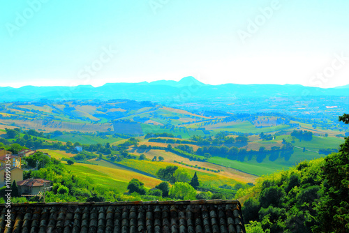 Magical view from Montelupone at a hazy valley full of vivid green nature strewn across placid Marche hills with a house roof in the foreground and the Sibillini mountains' outlines in the background photo