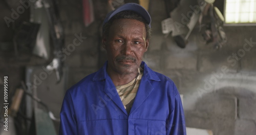 Portrait of a happy senior African male panel beater in a township workshop, wearing a cap, looking