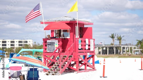 Vibrant lifeguard tower on a sunny beach with American and yellow flags waving, surrounded by beachgoers and colorful umbrellas. A tropical setting perfect for safety, summer, and tourism themes.