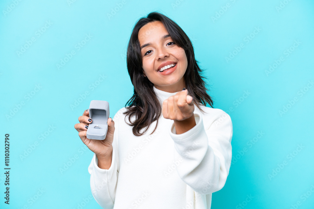 Young hispanic woman holding a engagement ring isolated on blue background making money gesture Young hispanic woman holding a engagement ring isolated on blue background making money gesture