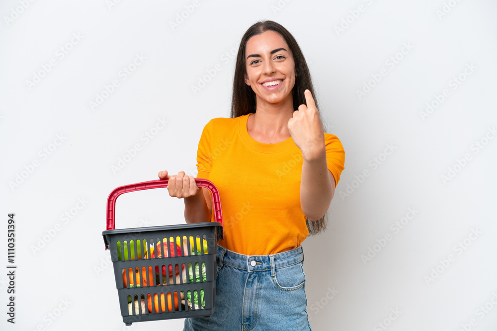 Young caucasian woman holding a shopping basket full of food isolated on white background doing coming gesture Young caucasian woman holding a shopping basket full of food isolated on white background doing coming gesture