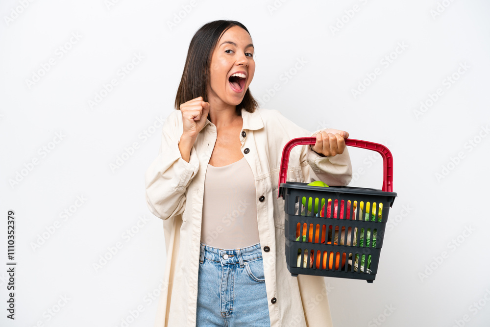 Young woman holding a shopping basket full of food isolated on white background celebrating a victory in winner position Young woman holding a shopping basket full of food isolated on white background celebrating a victory in winner position