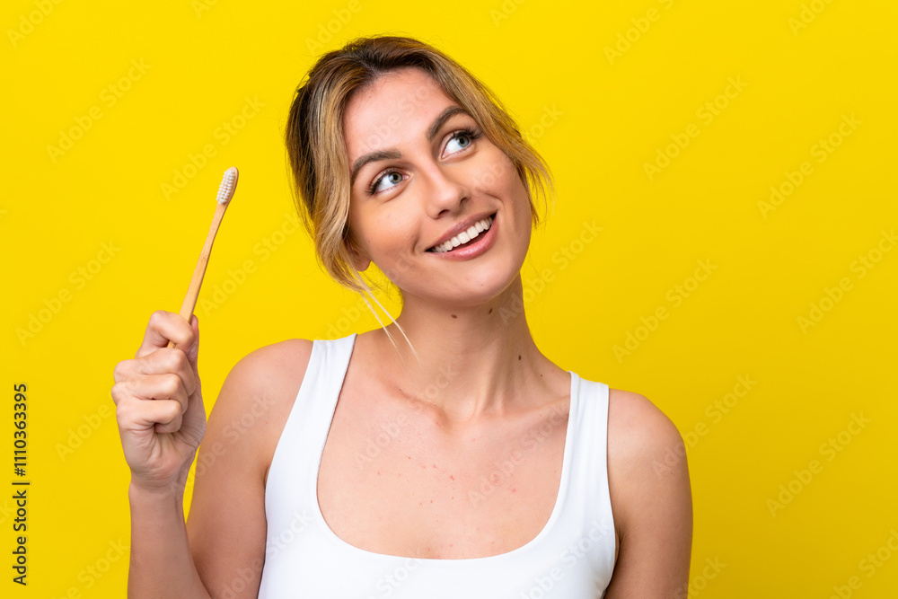 Young Uruguayan woman brushing teeth isolated on yellow background looking up while smiling