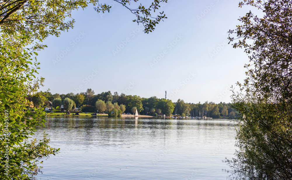 Obraz premium A serene lakeside view with a sandy beach, sailboats, and lush green trees under a clear sky, showcasing a peaceful outdoor recreation scene.