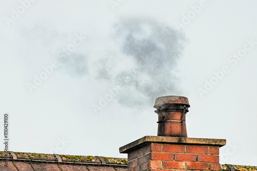 Smoke plume rising from brick chimney with bird guard cowl on mossy roof against cloudy sky