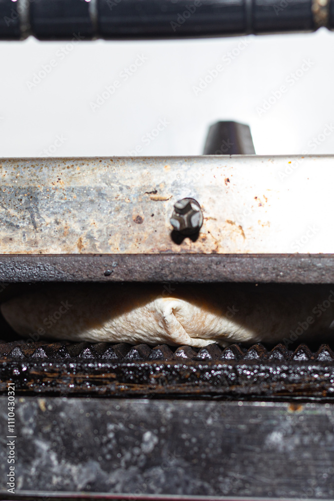 The lower view of a panini press in operation, cooking a wrap to perfection. The photograph emphasizes the raw and industrial feel of the equipment and the food preparation process.