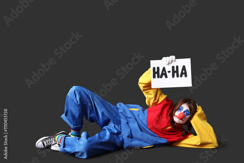 Young woman dressed as sad clown holding card with text HA-HA on black background. April Fool's day celebration