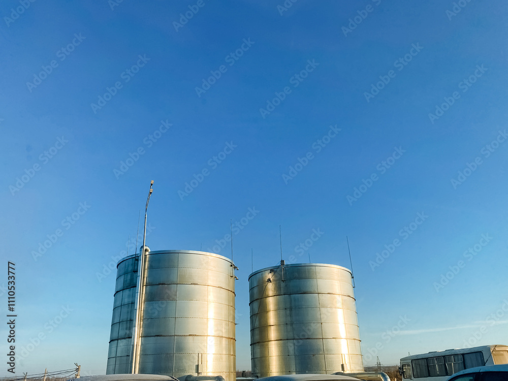 Two large, shiny stainless steel tanks stand tall against a vibrant blue sky, providing essential storage for various liquids, including gas, within an urban engineering landscape Two large, shiny stainless steel tanks stand tall against a vibrant blue sky, providing essential storage for various liquids, including gas, within an urban engineering landscape