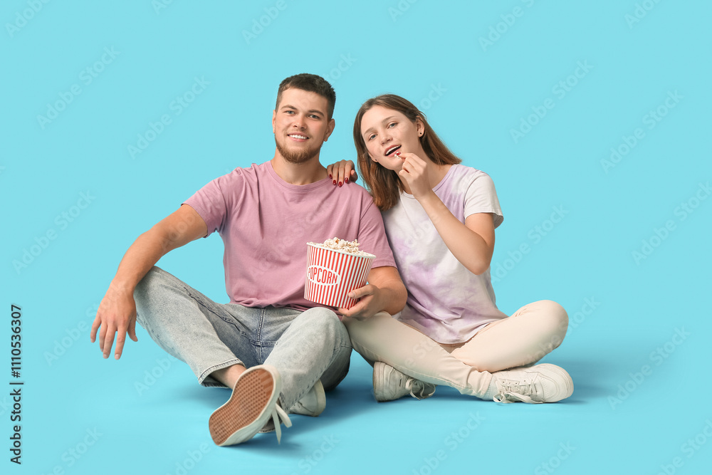 Happy young couple with bucket of popcorn sitting on blue background Happy young couple with bucket of popcorn sitting on blue background