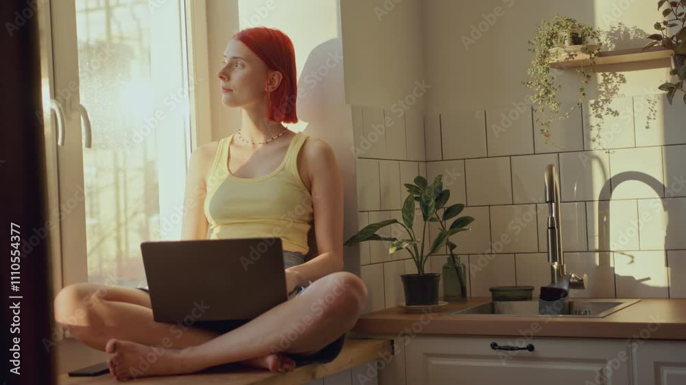 Young redhead girl sitting cross-legged on windowsill in sunlit kitchen, looking out and working on laptop during day at home Young redhead girl sitting cross-legged on windowsill in sunlit kitchen, looking out and working on laptop during day at home