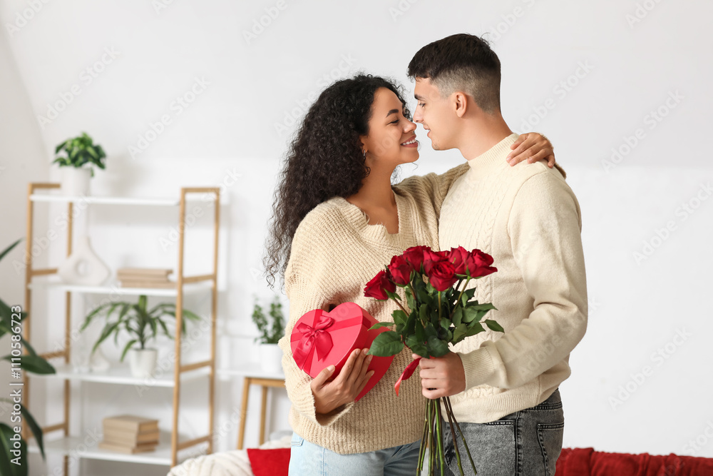 Loving young couple with gift box and bouquet of roses kissing at home. Valentine's Day celebration