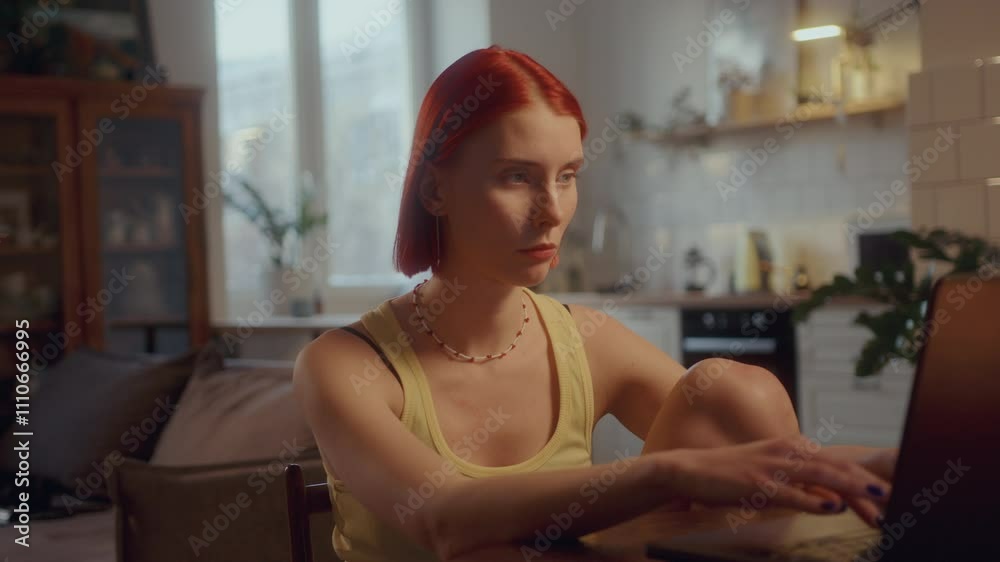 Pretty red-haired girl sitting at table in living room and typing on laptop, working remotely from home or communicating online Pretty red-haired girl sitting at table in living room and typing on laptop, working remotely from home or communicating online