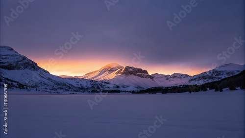 The sun's last rays illuminate the snow-clad peaks of a mountain range, highlighting the serene beauty of a wintery landscape under a dusky sky