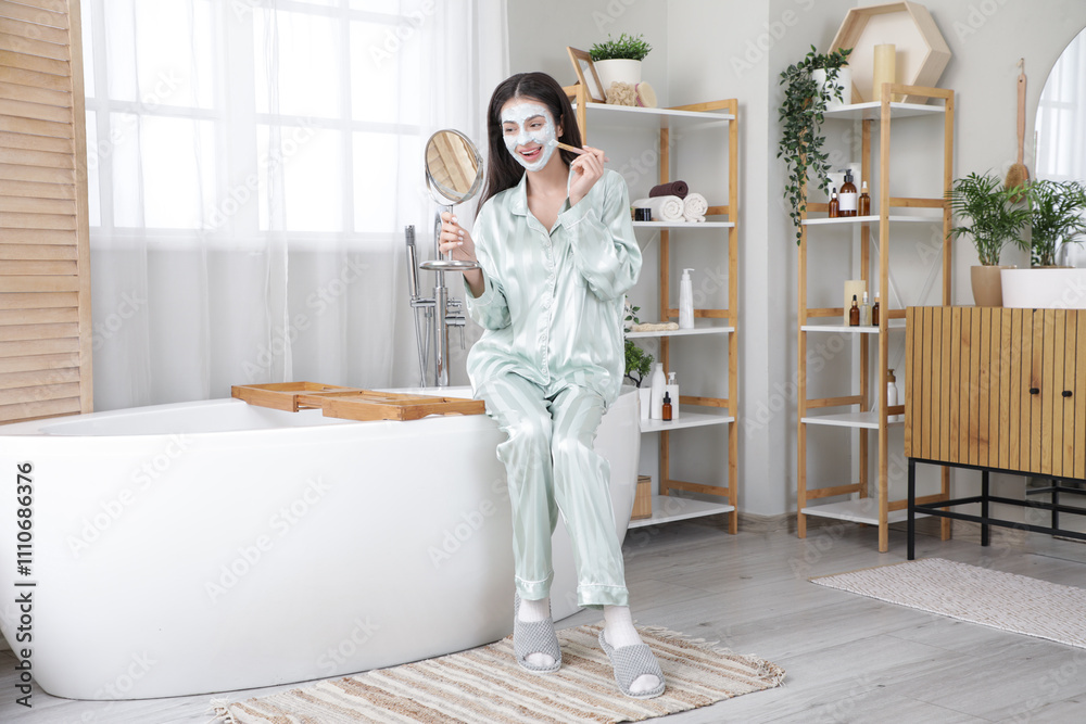 Beautiful young woman with mirror applying facial mask in bathroom Beautiful young woman with mirror applying facial mask in bathroom