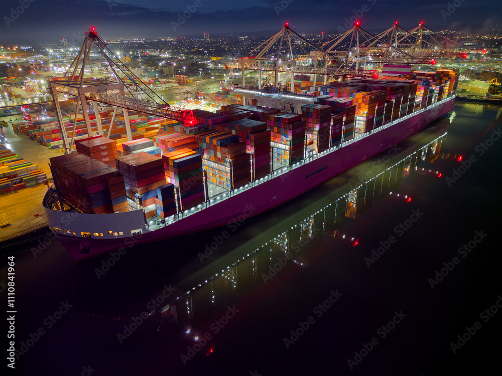 Side view of large container ship at illuminated commercial docks in Southampton UK. Clear lights reflection on calm sea. Night aerial view of single vessel in pink color moored at container terminal.