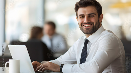 Smiling Businessman in Formal Shirt and Tie Working on Laptop in a Bright Modern Office Environment with Colleagues in Background