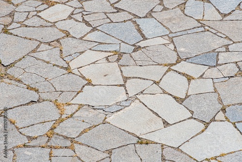 Close-up image of weathered, irregular stone tiles arranged in a herringbone pattern on a paved surface Muted earthy tones dominate the scene with some worn areas revealing underlying dirt or smalle photo