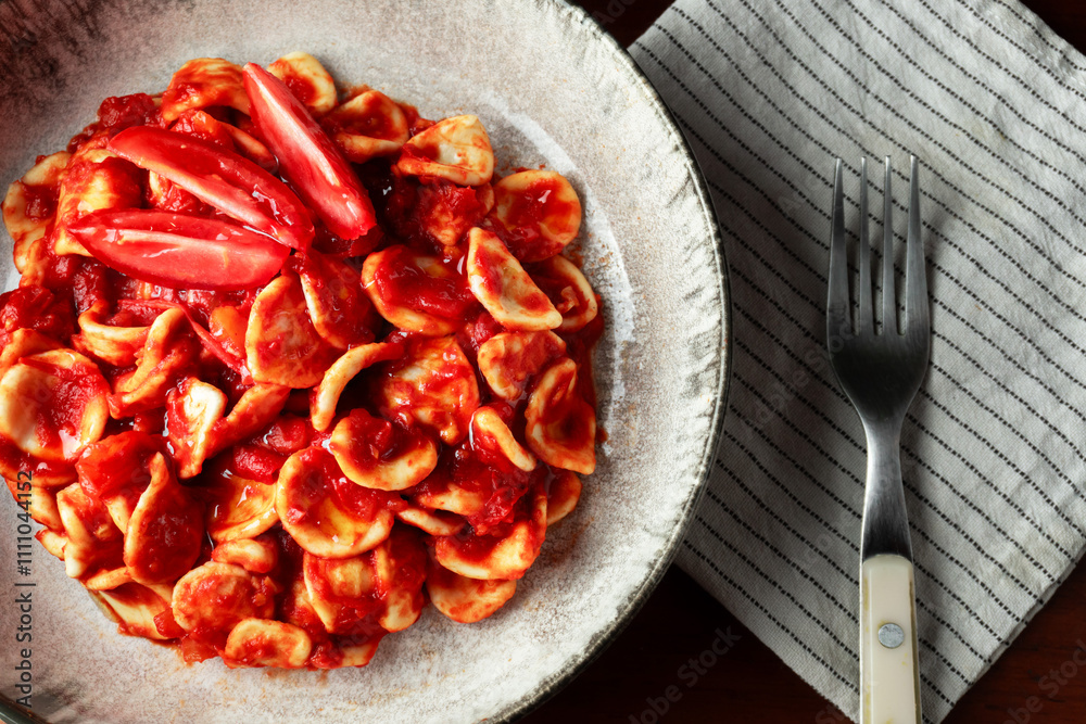 Tipical italian homecooked meal: orecchiette pasta with fresh tomato sauce. The dish is served in a vintage plate and placed next to a fork and a striped napkin. Dark, moody look, wooden table Tipical italian homecooked meal: orecchiette pasta with fresh tomato sauce. The dish is served in a vintage plate and placed next to a fork and a striped napkin. Dark, moody look, wooden table