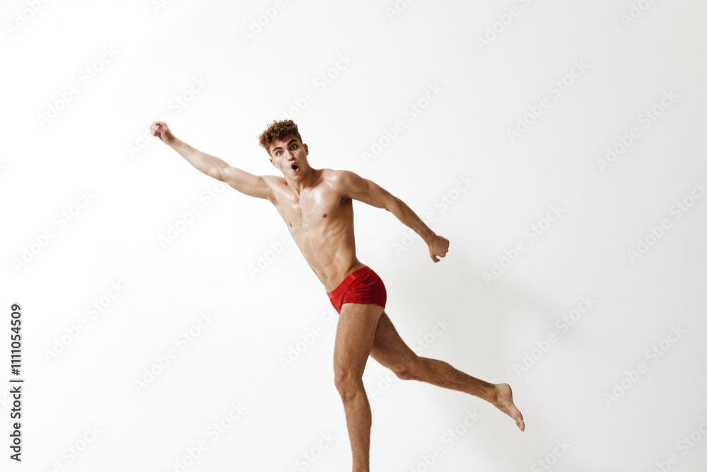 Obraz premium Energetic young man in red swimwear posing playfully, conveying joy and enthusiasm. Bright white background emphasizes movement and fitness.
