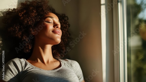 A serene image of a woman with curly hair, eyes closed, basking in warm sunlight inside a room, conveying calmness and contentment, emphasizing peace and relaxation.