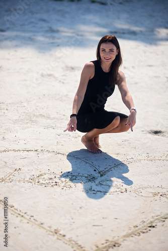 Woman in a black dress crouching on a sandy beach, pointing at heart-shaped drawing