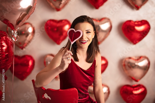 Joyful woman in a red dress holding a heart-shaped lollipop, surrounded by romantic balloons