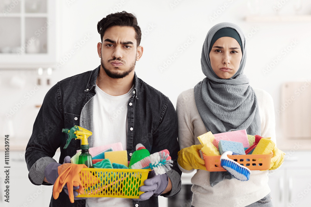Bored muslim family husband and wife cleaning their new apartment, holding house-keeping supplies, white kitchen interior, copy space. House-keeping service, apartment cleaning concept Bored muslim family husband and wife cleaning their new apartment, holding house-keeping supplies, white kitchen interior, copy space. House-keeping service, apartment cleaning concept