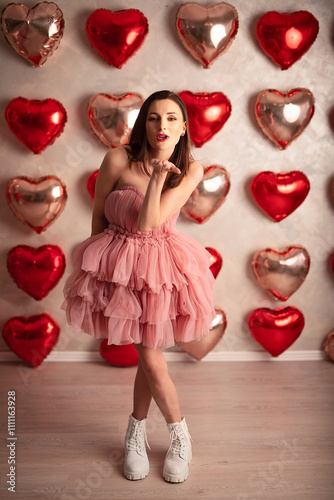 Playful woman in a pink ruffled dress blowing a kiss, surrounded by heart-shaped balloons