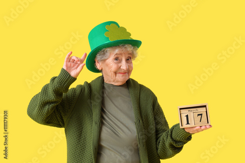 Senior woman in leprechaun hat with calendar for St. Patrick's Day celebration on yellow background photo