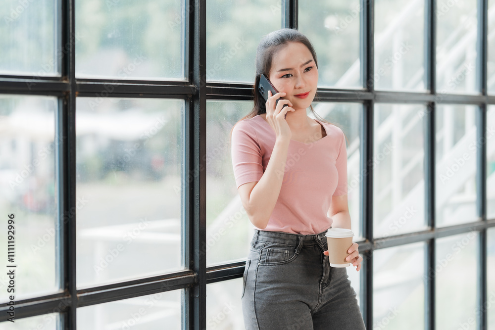 Modern Businesswoman on a Call: A stylish young Asian woman in a pink shirt and gray jeans stands by a large window, casually holding a coffee cup while engrossed in a phone call. Modern Businesswoman on a Call: A stylish young Asian woman in a pink shirt and gray jeans stands by a large window, casually holding a coffee cup while engrossed in a phone call.