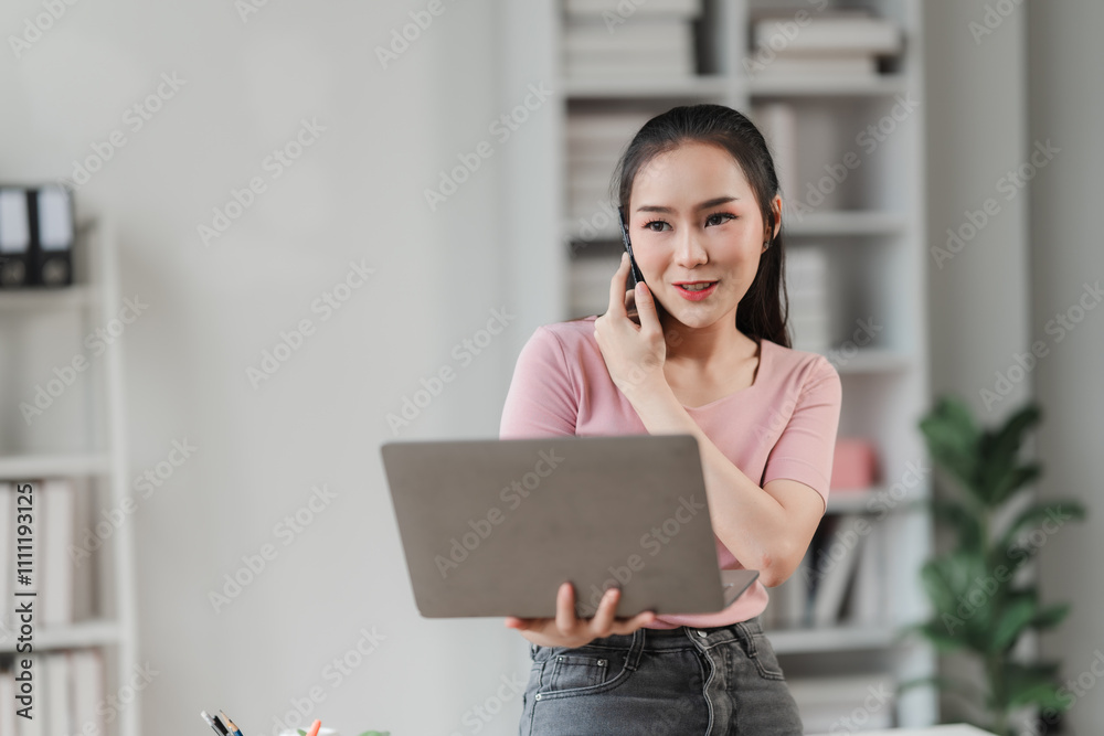 Confident Businesswoman on a Call: A young Asian businesswoman smiles confidently while working on her laptop and taking a phone call in her modern home office. Confident Businesswoman on a Call: A young Asian businesswoman smiles confidently while working on her laptop and taking a phone call in her modern home office.
