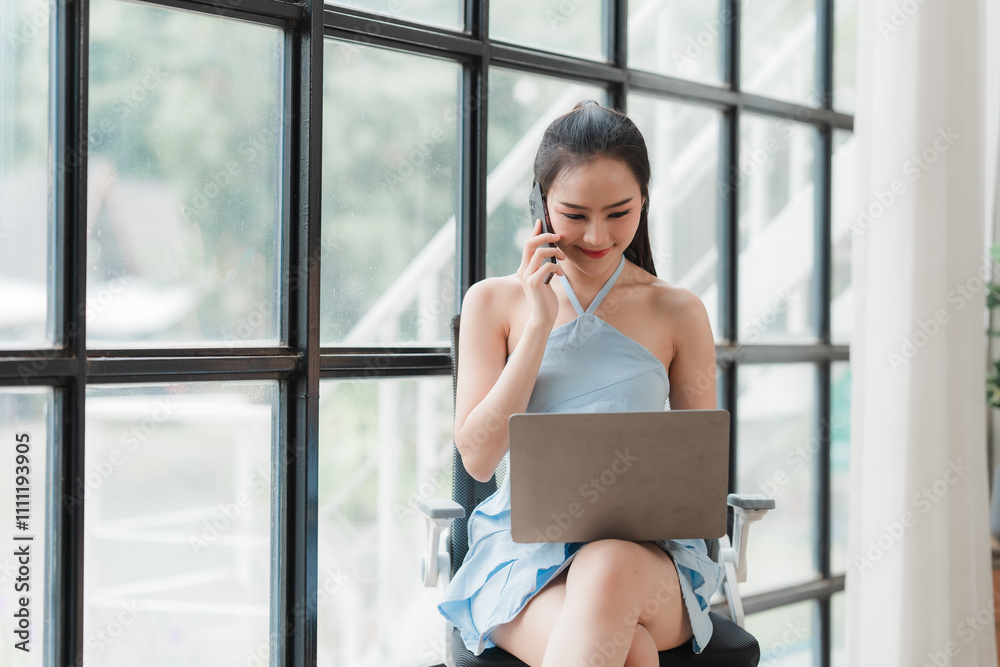 Confident Businesswoman: A young businesswoman in a blue dress sits by a large window, expertly navigating her laptop while on a phone call. Confident Businesswoman: A young businesswoman in a blue dress sits by a large window, expertly navigating her laptop while on a phone call.