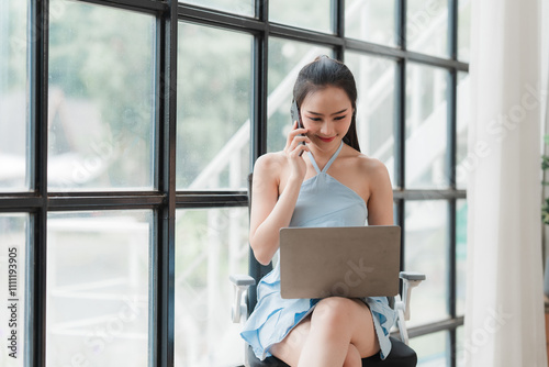 Confident Businesswoman: A young businesswoman in a blue dress sits by a large window, expertly navigating her laptop while on a phone call.