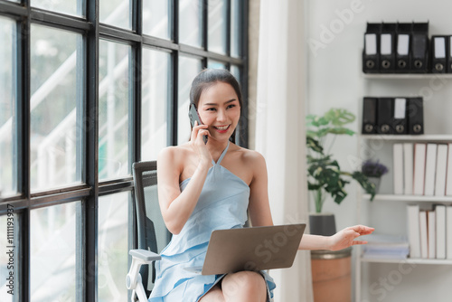 Businesswoman on a Call: A confident and professional woman in a blue dress, effortlessly multitasks by taking a phone call and working on her laptop in her modern office. photo