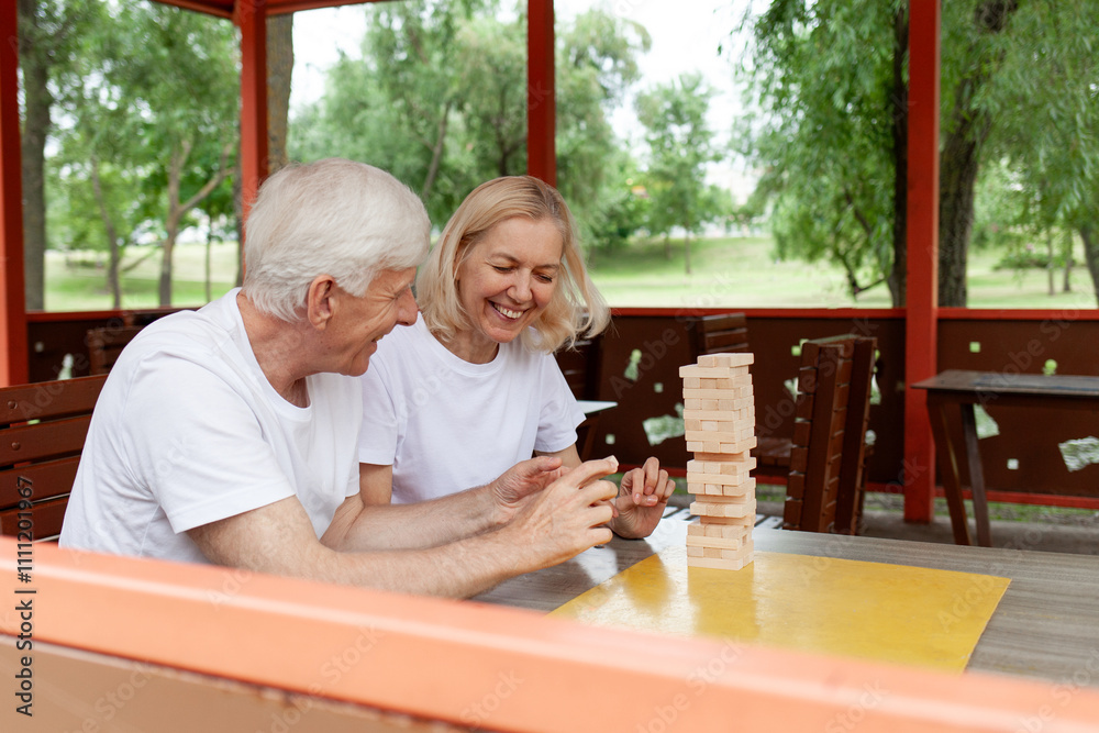 elderly couple of seniors in white t-shirts playing board game in park outdoors, old man and woman building a tower of wooden bricks and competing in a game of balance elderly couple of seniors in white t-shirts playing board game in park outdoors, old man and woman building a tower of wooden bricks and competing in a game of balance