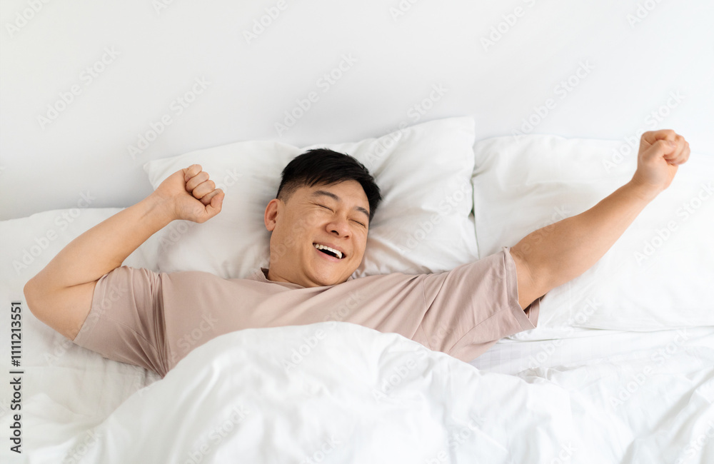 Top view of smiling asian middle aged man enjoying happy morning, lying in comfy bed with closed eyes at home, stretching his body after waking up, feeling excited and well-rested, copy space