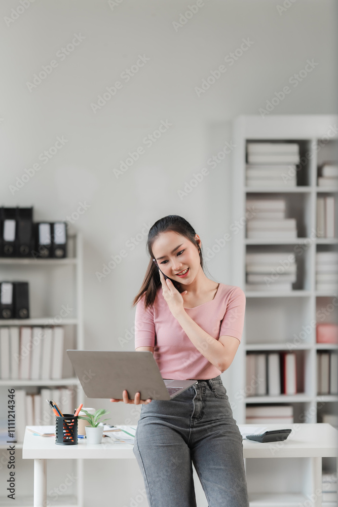 Focused and Connected: A young businesswoman leans against her desk, engrossed in a conversation on her phone while reviewing documents on her laptop. Focused and Connected: A young businesswoman leans against her desk, engrossed in a conversation on her phone while reviewing documents on her laptop.