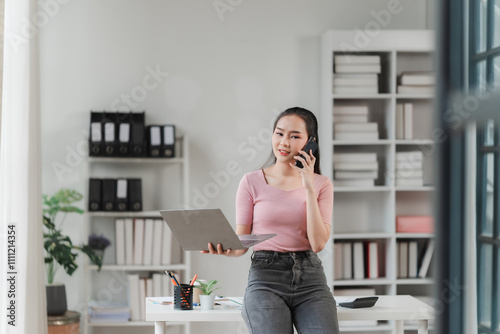 Focused and Connected: A young businesswoman multitasks effortlessly, balancing a phone call and her laptop in a modern, bright office setting. photo