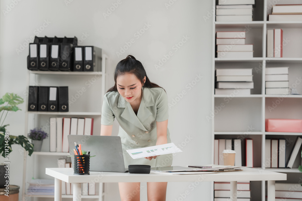 Businesswoman Analyzing Data: A focused young businesswoman examines a document and laptop in her modern office, demonstrating dedication and determination in the pursuit of professional success. Businesswoman Analyzing Data: A focused young businesswoman examines a document and laptop in her modern office, demonstrating dedication and determination in the pursuit of professional success.