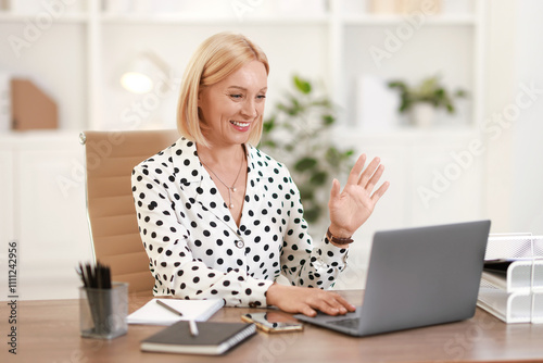 Smiling middle aged woman having videochat by laptop at table in office