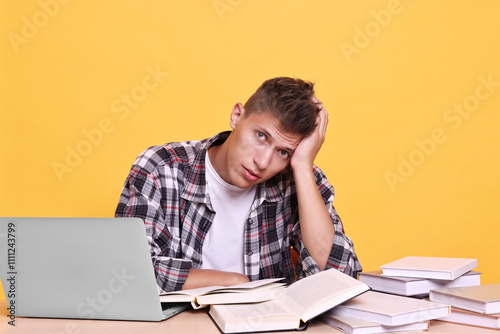 Young student with books and laptop having stress before exam at table against yellow background photo